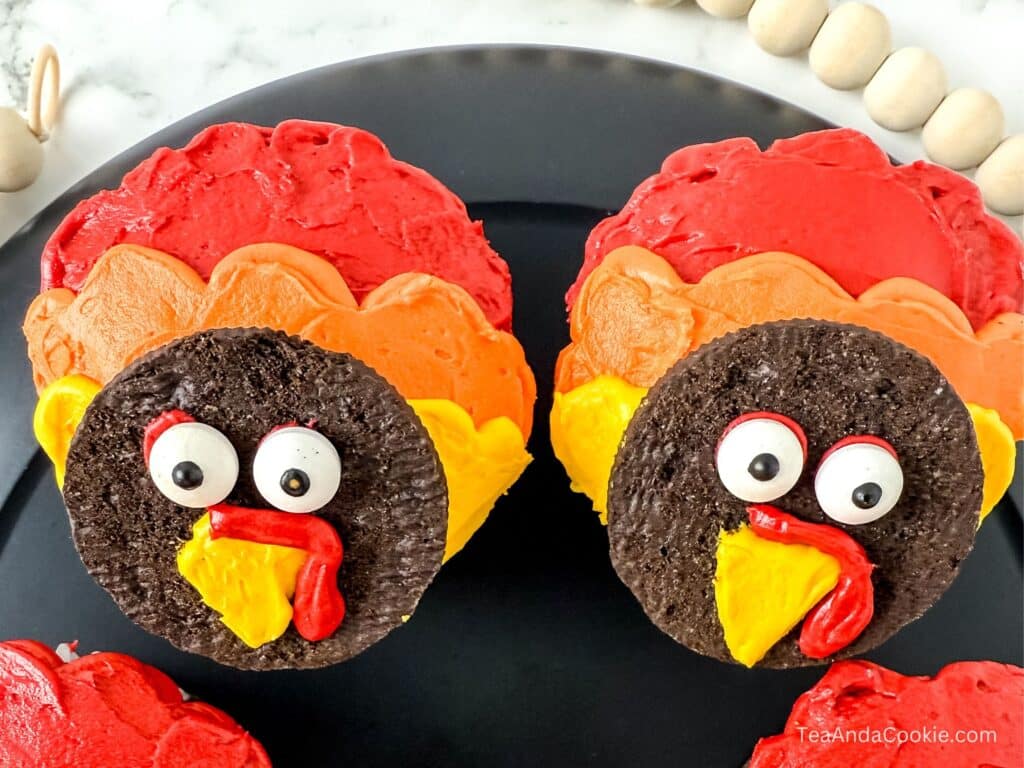 Turkey Cupcakes With Oreos. Two turkey-themed cookies with chocolate faces, candy eyes, and colorful red, orange, and yellow frosting feathers are displayed on a black plate.