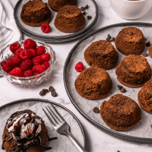 Three plates hold multiple chocolate lava cakes, with a bowl of fresh raspberries and a few chocolate chips nearby. One lava cake is topped with ice cream and chocolate sauce on a small plate with a fork.