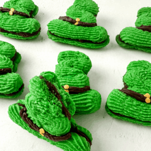 Green cookies shaped like leprechaun hats with chocolate bands and small yellow candy accents, arranged on a white surface.