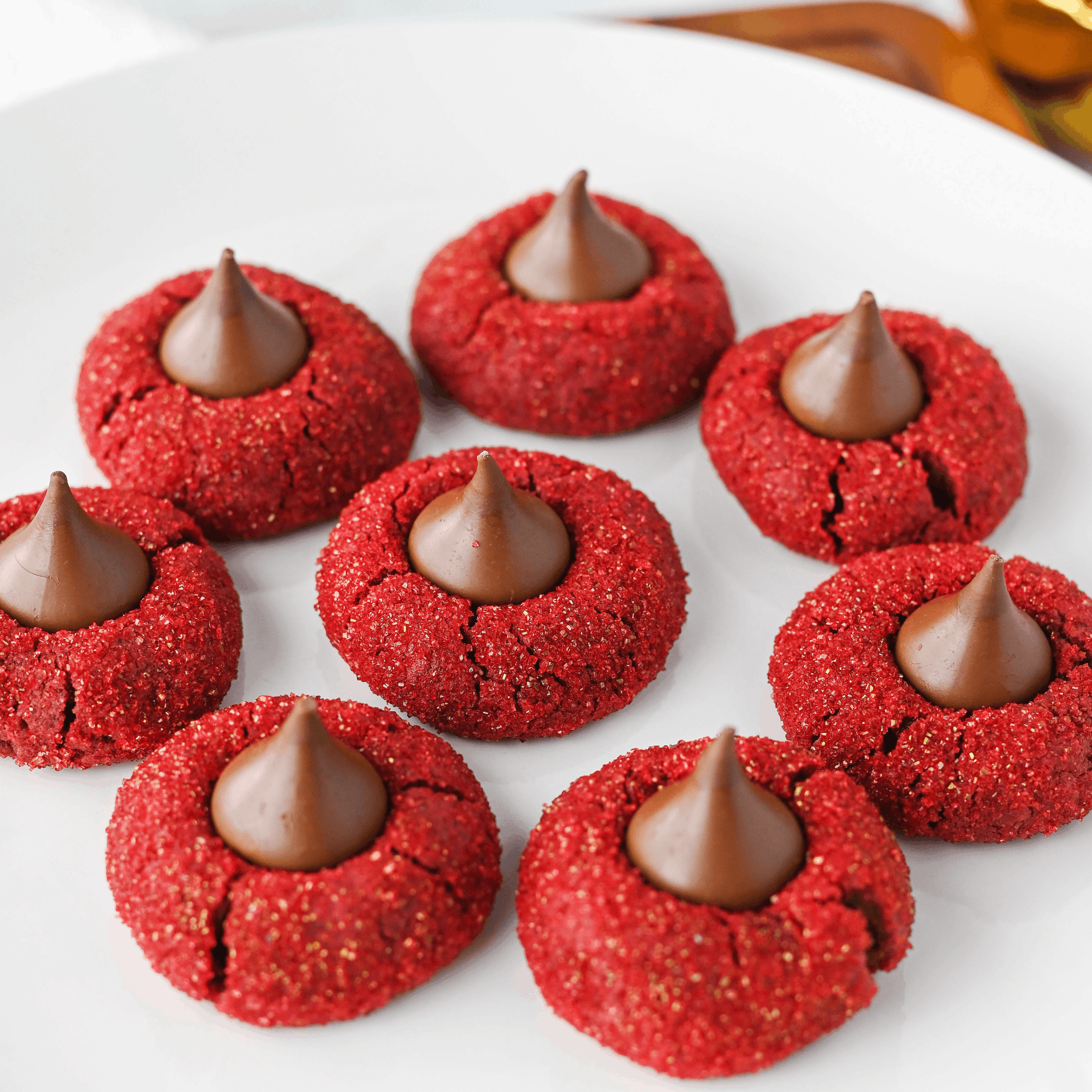 Red velvet blossom cookies. Seven red sugar cookies topped with chocolate kisses are arranged on a white plate.