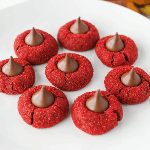 A white plate with seven red cookies, each topped with a chocolate kiss, arranged in a circular pattern.